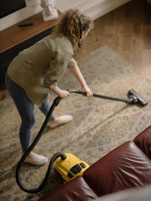 Close-up of a vacuum cleaner head actively cleaning an ornate, patterned carpet in a residential living room; the dark-colored vacuum attachment is pressed firmly against the light-colored, intricate
